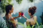 Zwei Frauen stoßen mit Getränken an einem Wasserufer an.